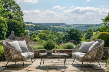 Fototapeta premium Light grey rattan garden furniture set on stone terrace with lush greenery and rolling hills.