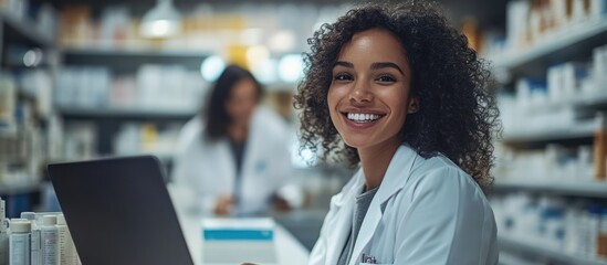 A smiling female pharmacist with curly hair in a white coat stands in front of a pharmacy counter with a laptop, and a colleague is in the background.