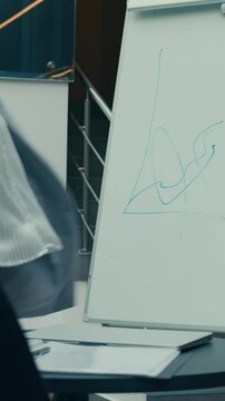 Vertical shot of female office worker in striped grey suit drawing graph on flipchart then starting making bizarre dance moves during business presentation in modern office space