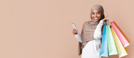 Cheerful afro muslim girl in hijab posing with smartphone and bright shopping bags over yellow...