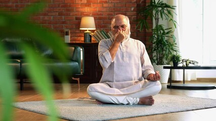 An Indian Asian elderly businessman with a beard is sitting comfortably on the floor of his modern home, wearing traditional Indian clothes and doing breathing exercises for health and  mental peace