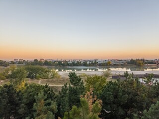 Fototapeta premium evening quiet city landscape with pine trees in the foreground, calm Kuban river and smoke from fires over trees on the other bank South of Russia, Krasnodar