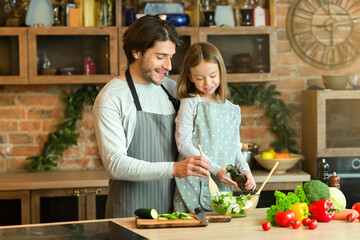 Cheerful young dad and little daughter preparing healthy salad in kitchen together, cooking lunch ant home, free space