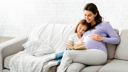 Obraz premium Happy family pregnant mother and preschooler daughter spending time together at home, reading book