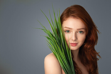 Naklejka premium Pretty Ginger-Haired Girl Posing With Green Plant Leaves Standing Looking At Camera Over Gray Studio Background. Natural Beauty, Spa And Skincare Cosmetics Concept