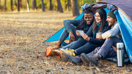 Smiling multiethnic backpackers looking at smartphone, drinking tea, camping together at autumn forest, empty space