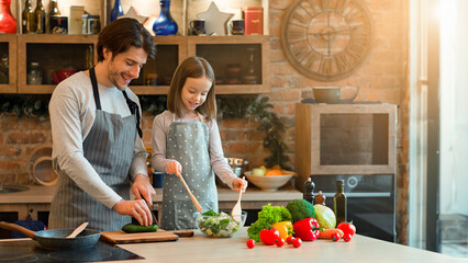 Little girl and her dad cooking healthy dinner together in kitchen at home, preparing vegetable salad for lunch, mixing ingredients, enjoying vegan organic homemade food, wearing aprons, copy space