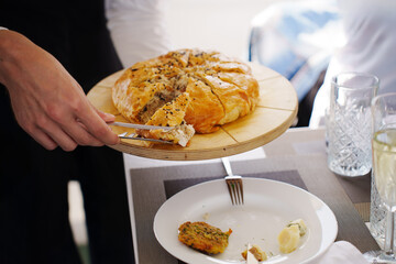 The waiter puts a piece of puff cake with meat on a plate. Traditional Russian pancake pie kurnik. © Maria