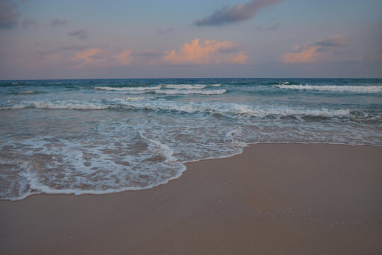 Sandy beach with gentle waves rolling in at sunset. The sky is a soft pink and purple, with fluffy clouds