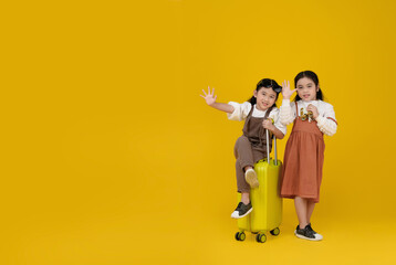 Two Asian smiling girls kids with a yellow suitcase, one waving energetically, and the other holding binoculars, standing against a bright yellow background with copy space.