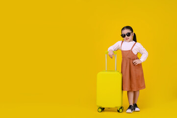 Asian confident girl kids wearing sunglasses and a brown dress stands confidently with a yellow suitcase, striking a pose against a bright yellow background.