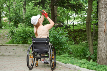 A woman in a cap using a wheelchair takes a photo with her phone outdoors.