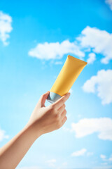 A mockup yellow cream tube is being held by a female model against a clear blue sky with white...