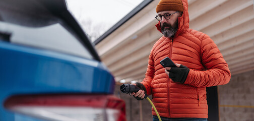 Fototapeta premium Man charging electric car during cold snowy day. Close up of man putting charger in charging port. Charging and driving electric vehicles during winter season.