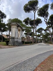 A  peaceful daytime scene in Villa Borghese, Rome, Italy, featuring a paved path lined with tall pine trees and classical stone monuments. The lush greenery and open space create a serene atmosphere i