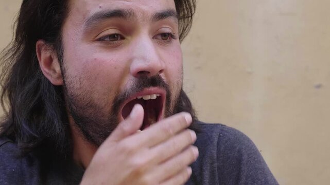 A young man with long hair and a short beard yawns, expressing tiredness and the need for rest. His relaxed demeanor is captured in a candid moment.