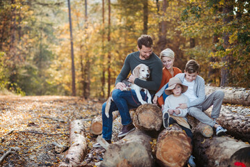 Portrait of family sitting on logs in the middle autumn forest. Nuclear family with dog spending weekend outdoors.