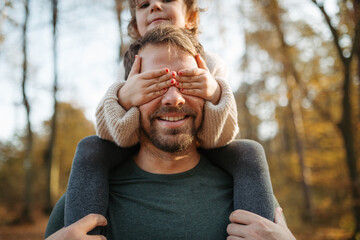 Father walking with daughter on his shoulders across autumn forest. Dad and girl having fun...