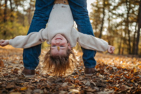 Father holding little girl by legs heand down, playing during walk in autumn forest. Dad and girl having fun outdoors.