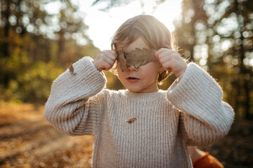 Portrait of cute girl standing in the middle of autumn forest, covering her eyes with dry leaves.