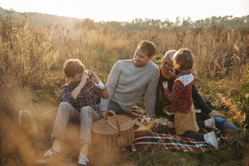 Portrait of family having picnic in autumn nature, eating sandwiches, fruits and cheese in the middle of meadow. Young boy taking pictures with vintage camera.