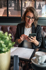Young attractive female sitting in a cafe, drinking coffee and looking at her phone. Young influencer preparing content for social media. Business woman smiling and looking at her smartphone.