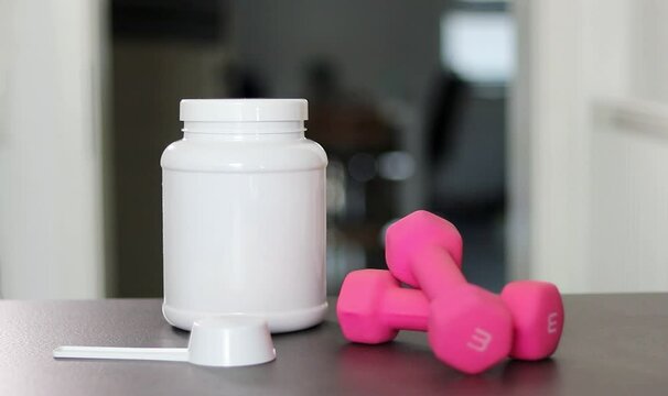 Close up of a protein powder box bottle on table with pink dumbbells and white whey protein powder in a scoop. Diet meal replacement for weight loss. 