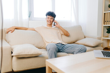 young man using smartphone while sitting on a comfortable sofa in a bright living room with natural light and soft decor, relaxed atmosphere, smiling joyfully, home comfort