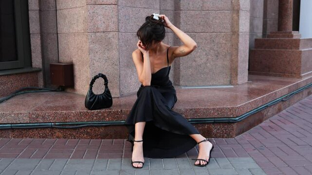 A mulatto girl in a black dress sits on the doorstep of a building in the city and looks at the camera. Businesswoman. The girl in Paris