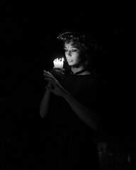 Fototapeta premium A witch in a dark forest, she is in a black dress. The witch has a wreath on her head. A beautiful young girl is dressed somberly, performs a ritual and practices magic