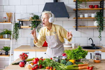 happy beautiful elderly gray haired senior woman in headphones cook in cozy kitchen with fresh organic vegetables, having fun and singing