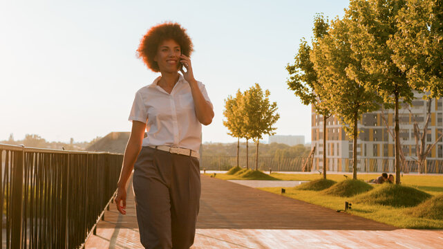 Happy smiling ethnic African American curly woman talking mobile phone smile walking outdoors business girl make online order smartphone talk call conversation businesswoman walk in city sun sunshine