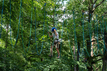 Teenager in a blue helmet navigates a challenging ropes course high above the ground in a dense...