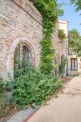 Fototapeta premium Stone Facade With Vines And Flowers On A Rustic House. The Arched Windows With Bars And The Surroundings Full Of Vegetation Create A Picturesque And Natural Atmosphere.