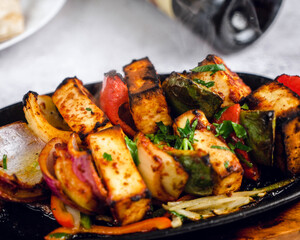 Spicy sizzling platter of colorful vegetable skewers, grilled to perfection, sits atop a wooden board. Accompanying the dish are soft naan bread and a bottle of rich red wine in the background.