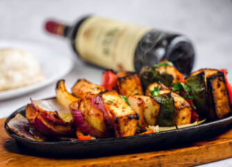 A sizzling platter of colorful vegetable skewers, grilled to perfection, sits atop a wooden board. Accompanying the dish are soft naan bread and a bottle of rich red wine in the background.