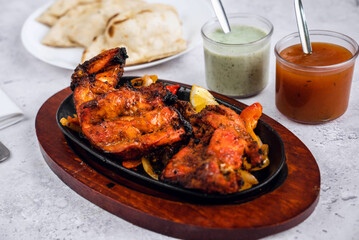 A vibrant, sizzling platter of tandoori chicken served on a wooden board, garnished with lemon slices. In the background, fluffy naan bread sits on a plate, accompanied by two dipping sauces in jars .