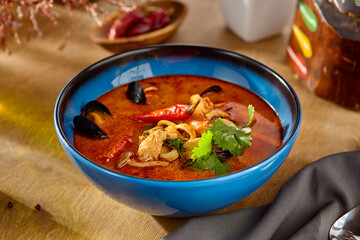 Tom Yum Soup with Seafood in Blue Bowl on Wooden Table Garnished with Cilantro and Chili Peppers