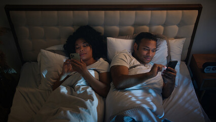 African American couple in bed at night late time in game bedroom relaxing using two phones chatting in mobile app scrolling social media smartphones woman man wife husband talking discuss news family