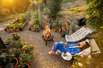 A woman enjoys a peaceful evening by the fire in a lush garden, surrounded by flowers and greenery....
