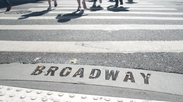 Broadway street sign, people on pedestrian zebra crossroad. New York City Manhattan road intersection. Text on asphalt near crosswalk, shadows on crossroad ped crossing. Traffic signage, United States