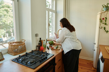 A confident woman happily chopping vegetables by the window.