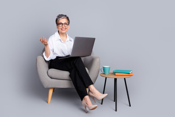 Full length photo of lovely senior lady sit armchair netbook dressed stylish formalwear isolated on gray color background © deagreez