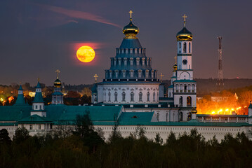 Golden full moon rising over the illuminated New Jerusalem Monastery in Istra, Russia, highlighting the Orthodox domes and towers in a tranquil evening landscape surrounded by trees