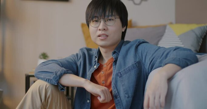 Slow motion of emotionless Asian guy sitting on floor in bedroom at home and looking at camera. Young man and accommodation concept.