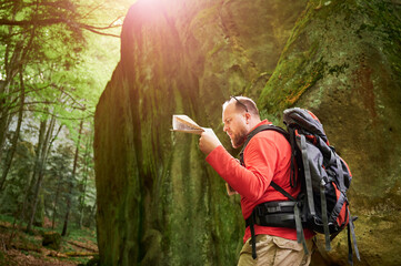 Bearded tourist man looks puzzled, studies map while stands in front of large rock in dense forest. Traveler with grey backpack and glasses, trying to figure out his direction.