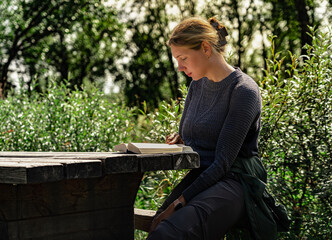 Fototapeta premium Beautiful woman sits quietly at a wooden table, engaged in a book while surrounded by greenery in a peaceful outdoor setting, enjoying reading in a serene moment in summer nature