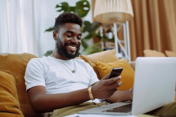 Fototapeta premium Man relaxes on couch with phone and laptop. Casual attire, white t-shirt, beige pants. Orange pillows, lamp in background. Warm glow, relaxed state, leisure, informality.