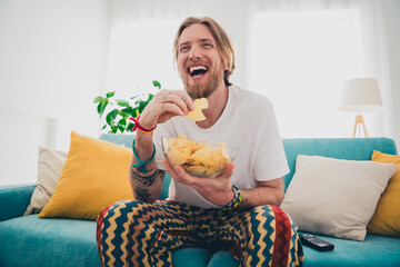 Photo of handsome young guy sit sofa watch tv laugh eat chips dressed nightwear spend pastime...