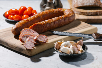 Smoked homemade sausage ring on the wooden cutting board with bowl of garlic, bread, tomato and knife. Rustic style. Hard light.